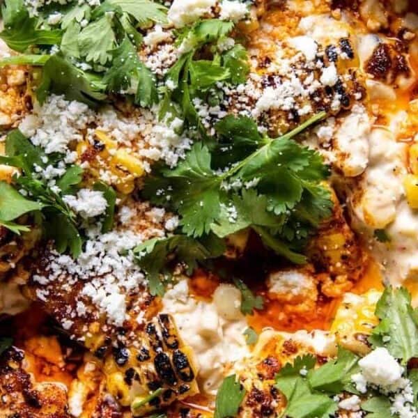 A bowl filled with creamy dip, topped with grilled corn, crumbled cheese, cilantro leaves, and a drizzle of sauce.