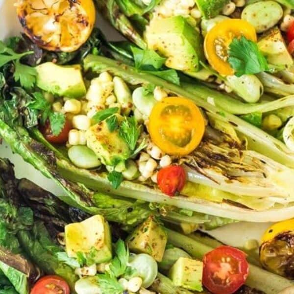Halved grilled romaine lettuce, avocado slices, corn kernels, cherry tomato halves, fresh herbs, and a grilled lemon half on a white plate.