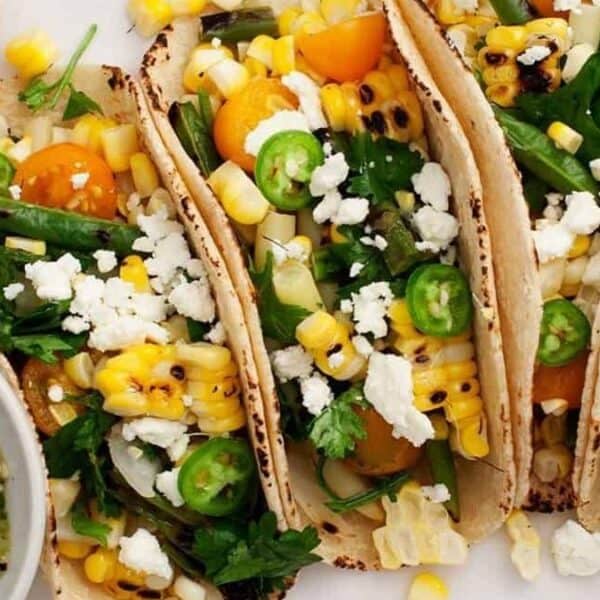 Three tacos filled with grilled corn, cherry tomatoes, jalapeño slices, fresh cilantro, and crumbled cheese are served with a side of salsa and lime wedges.