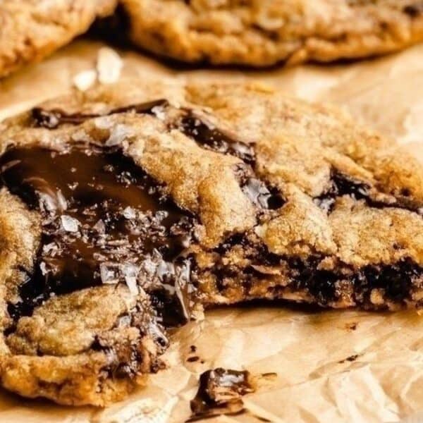 A chocolate chip cookie with a bite taken out sits on brown parchment paper. Melted chocolate is visible in the cookie, and additional cookies are blurred in the background.