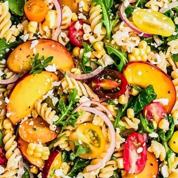 A bowl containing rotini pasta, sliced peaches, cherry tomatoes, corn kernels, arugula leaves, red onion slices, and crumbled cheese. A wooden spoon rests alongside.