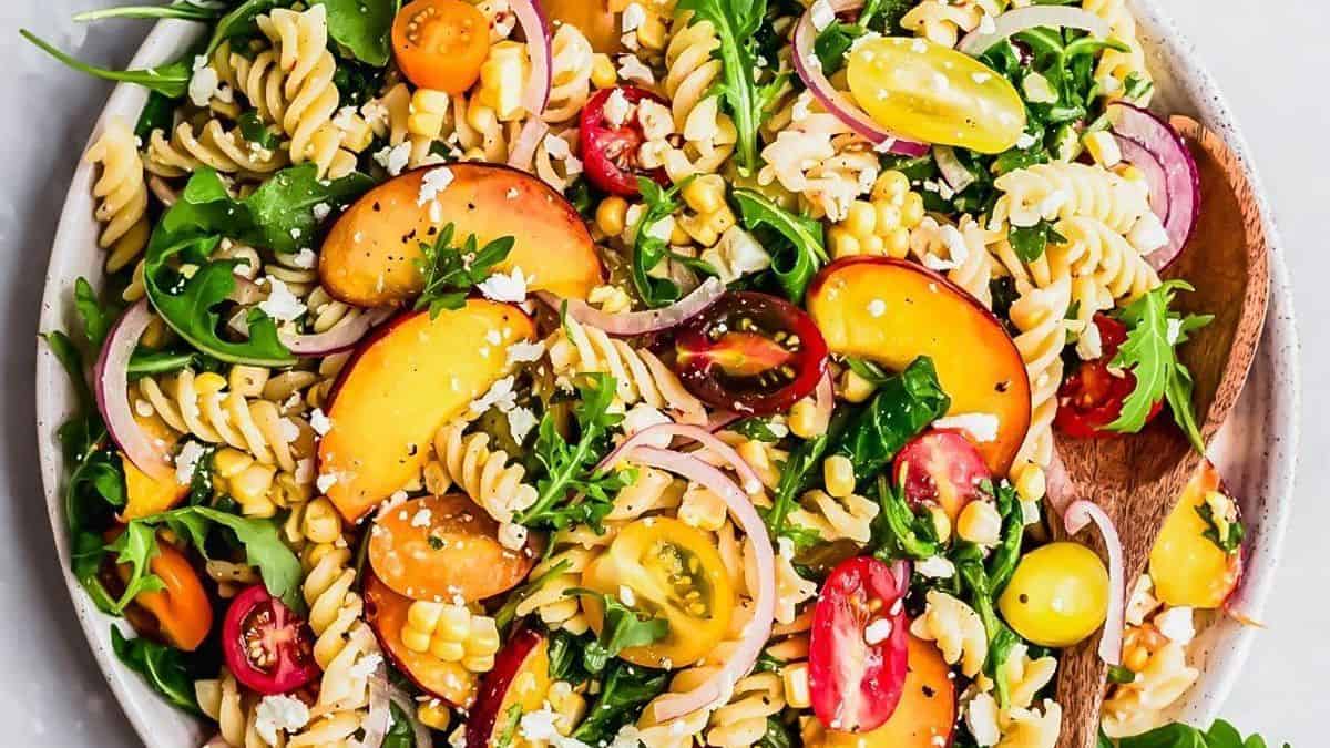 A bowl containing rotini pasta, sliced peaches, cherry tomatoes, corn kernels, arugula leaves, red onion slices, and crumbled cheese. A wooden spoon rests alongside.