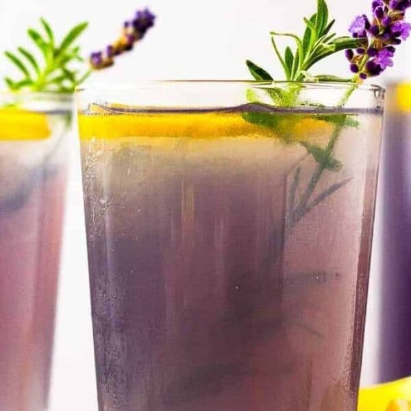 Three glasses filled with iced lavender lemonade, each garnished with a lemon slice, a sprig of lavender, and a sprig of rosemary, set against a light background.