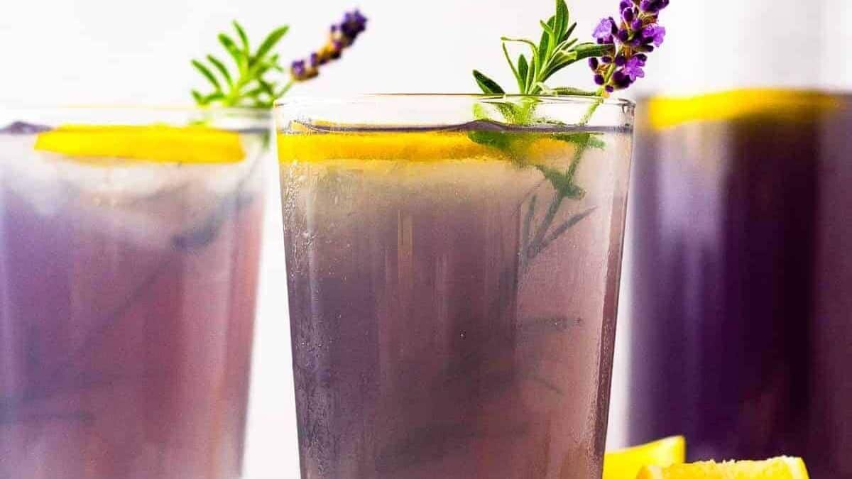 Three glasses filled with iced lavender lemonade, each garnished with a lemon slice, a sprig of lavender, and a sprig of rosemary, set against a light background.