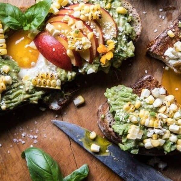 Sliced toast topped with mashed avocado, grilled corn, peach slices, a soft-cooked egg, fresh basil leaves, edible flowers, and a sprinkling of sea salt on a wooden board beside a knife.