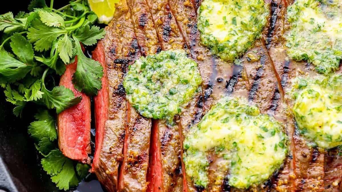 A grilled steak with a dollop of herb butter on top, accompanied by fresh cilantro and a lemon wedge.