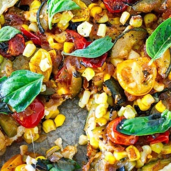 A vegetable galette with a golden crust, topped with sliced tomatoes, corn kernels, basil leaves, and assorted vegetables.
