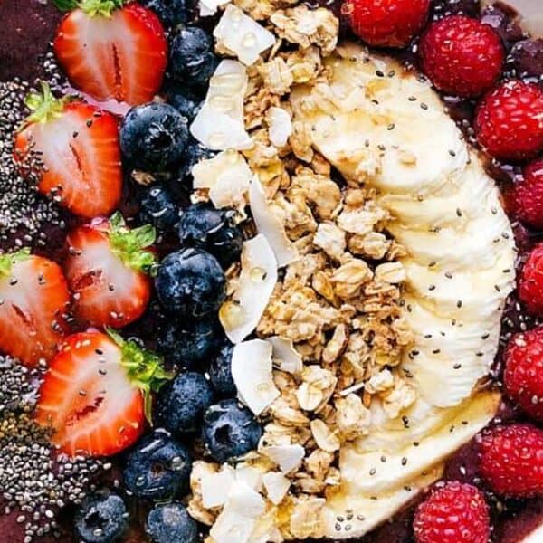 A bowl containing an acai base, topped with sliced strawberries, blueberries, banana slices, raspberries, granola clusters, coconut flakes, and chia seeds. A spoon is placed on the side.