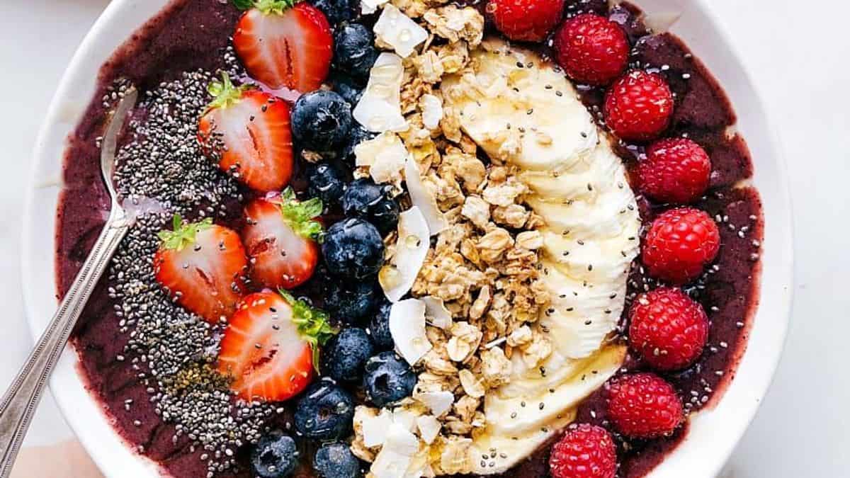 A bowl containing an acai base, topped with sliced strawberries, blueberries, banana slices, raspberries, granola clusters, coconut flakes, and chia seeds. A spoon is placed on the side.