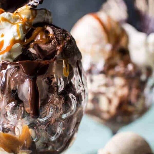 A glass dessert cup filled with scoops of chocolate and vanilla ice cream, topped with whipped cream, drizzled with caramel and chocolate sauce. In the background, two more dessert cups appear slightly blurred.