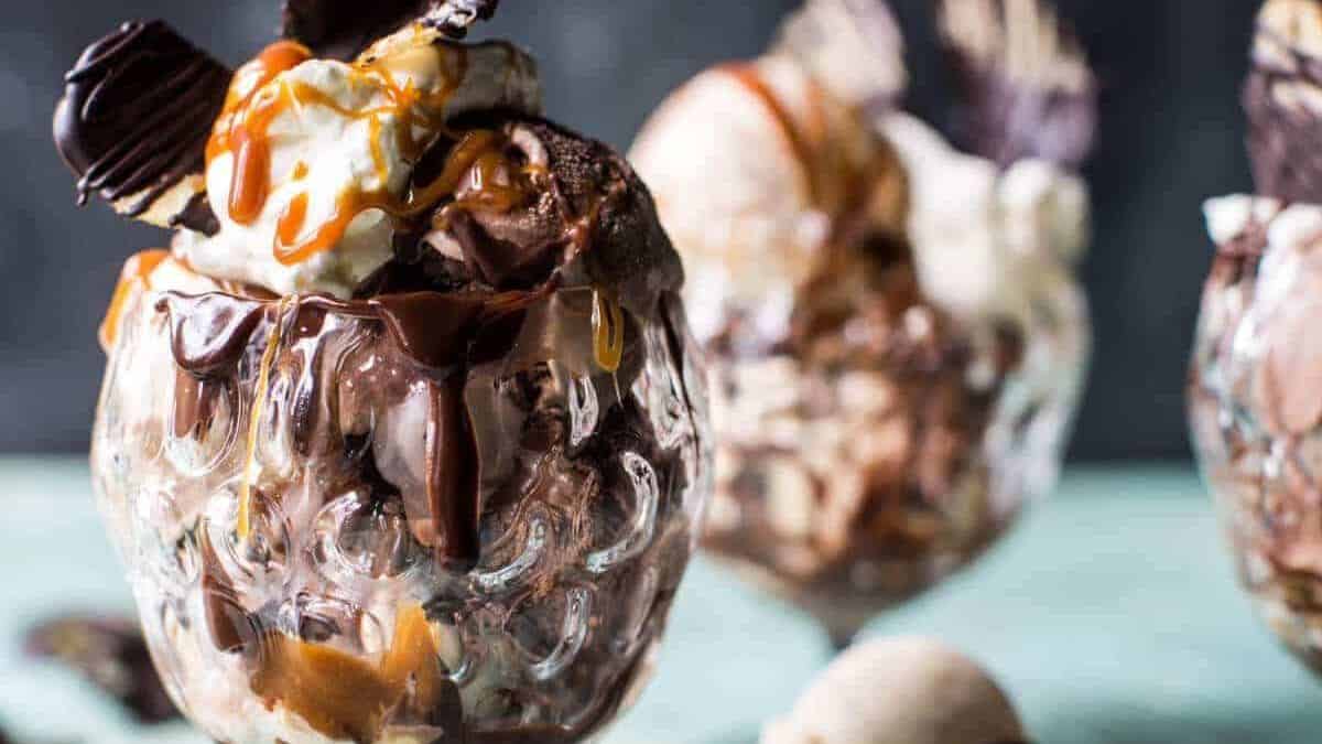A glass dessert cup filled with scoops of chocolate and vanilla ice cream, topped with whipped cream, drizzled with caramel and chocolate sauce. In the background, two more dessert cups appear slightly blurred.