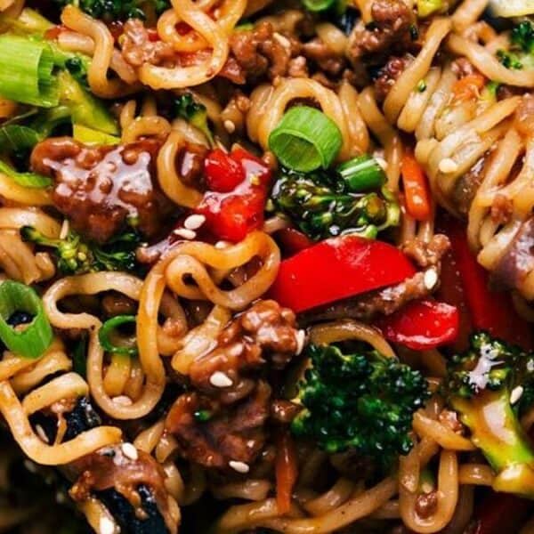 Stir-fried noodles topped with ground meat, broccoli florets, sliced red bell peppers, carrot strips, green onions, and sprinkled sesame seeds; a fork holds a portion of the noodles.