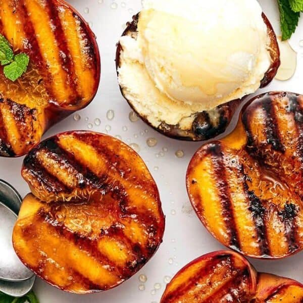 Grilled peach halves are on a tray, each topped with a scoop of vanilla ice cream and a sprig of mint. A spoon rests beside them.