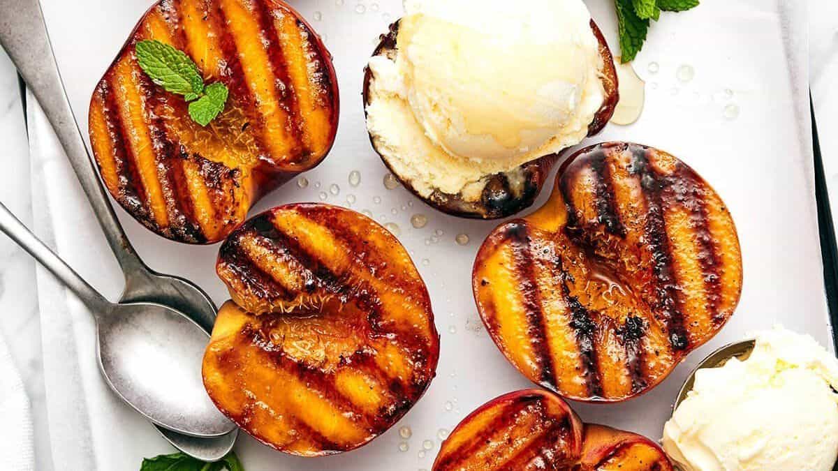 Grilled peach halves are on a tray, each topped with a scoop of vanilla ice cream and a sprig of mint. A spoon rests beside them.