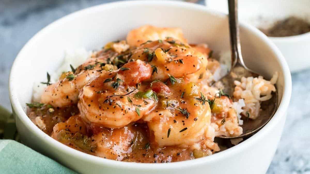 A bowl filled with shrimp étouffée over white rice, topped with chopped herbs, and a spoon resting inside the bowl.