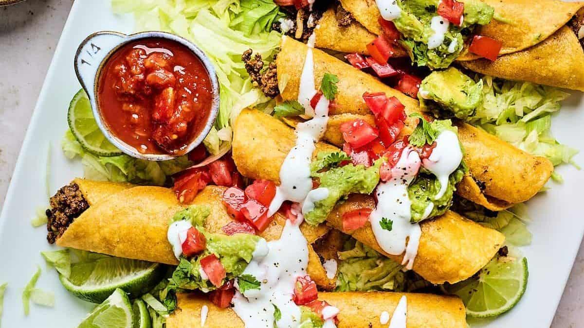 A plate of crispy taquitos topped with guacamole, diced tomatoes, cilantro, and sour cream, served with shredded lettuce, lime wedges, and a small bowl of salsa.