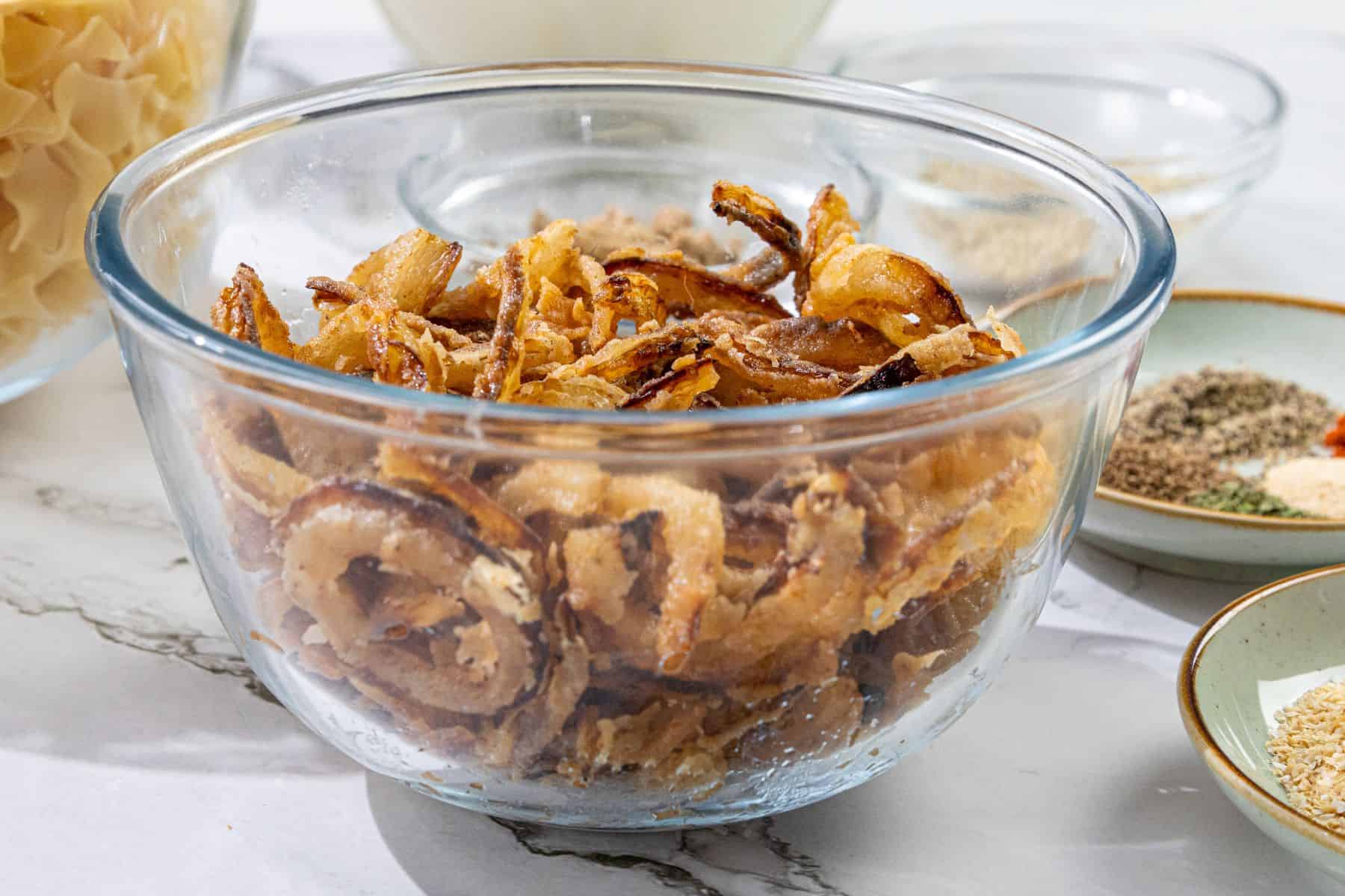 A clear glass bowl holds crispy fried onion strings on a marble countertop. Nearby are small dishes containing various spices and uncooked pasta.