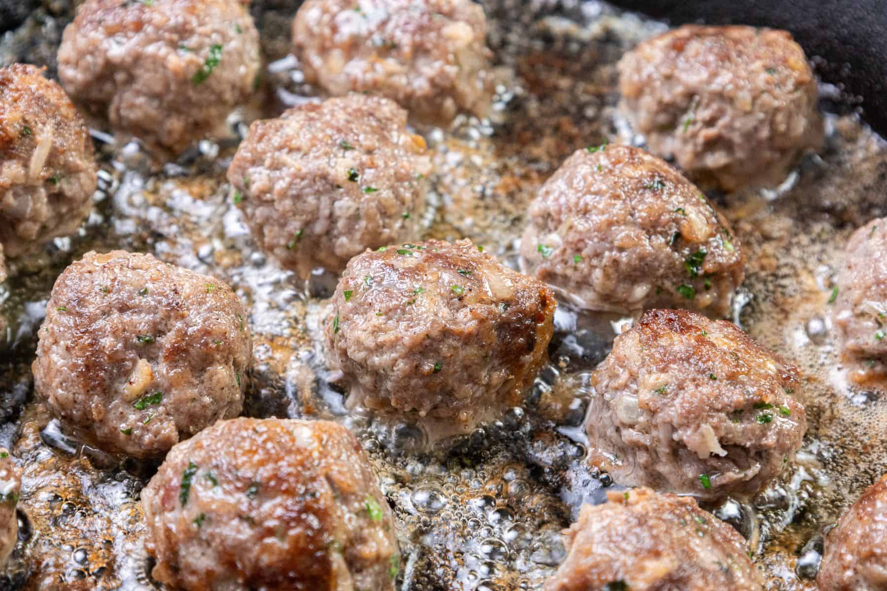 Several meatballs browning in a frying pan with hot oil.