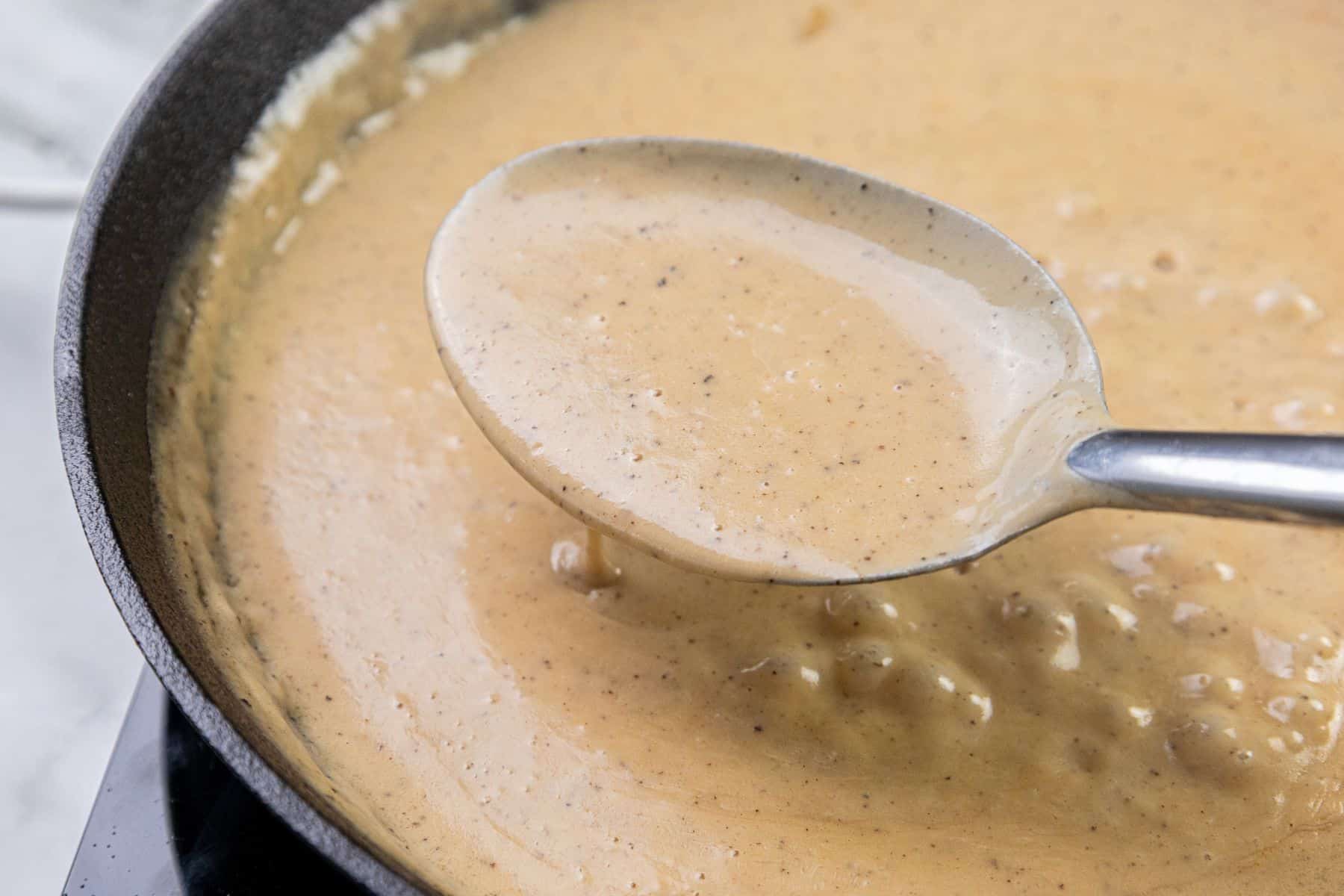 A spoon holds creamy, light brown gravy with visible pepper specks above a pan.