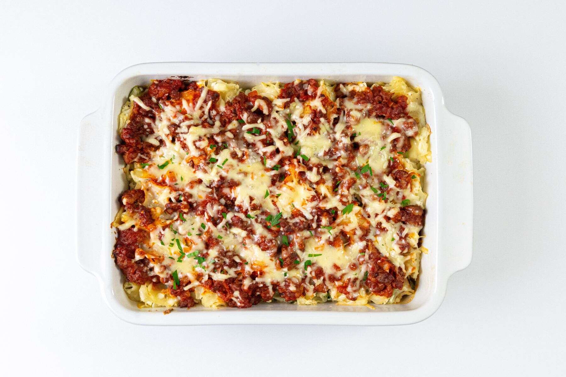 A white rectangular dish filled with lasagna, topped with golden melted cheese, visible tomato sauce, ground meat, and sprinkled chopped herbs, set against a white background.