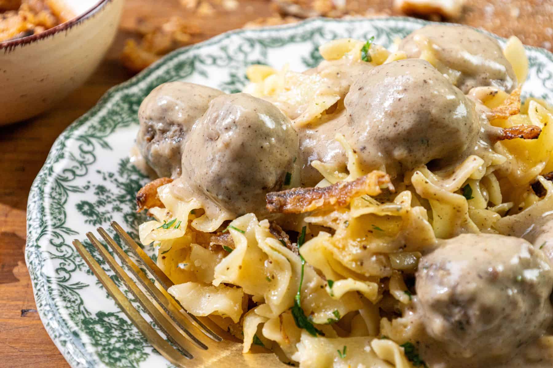 Egg noodles topped with meatballs in creamy gravy, garnished with herbs and crispy onions, served on a plate with a fork on the side.