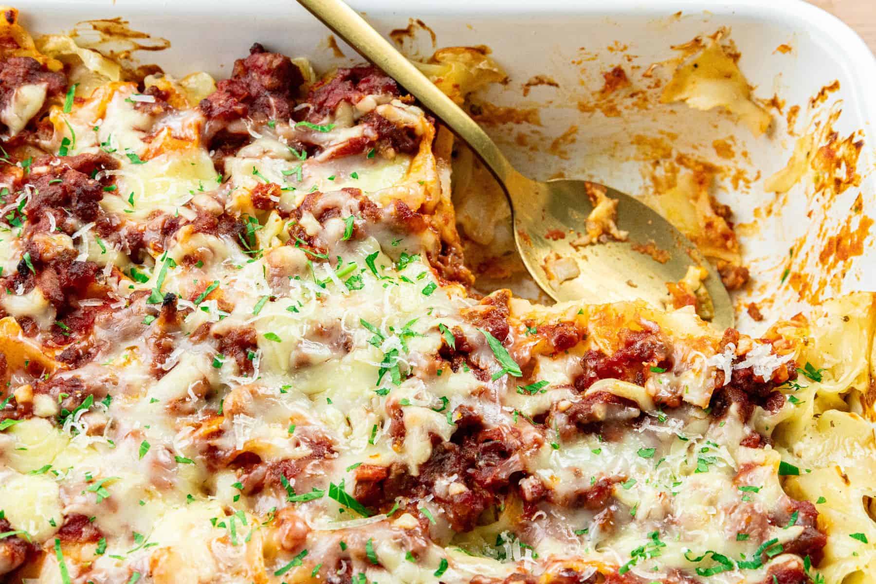 Baked pasta casserole with melted cheese, tomato sauce, and herbs in a white dish with a gold serving spoon.