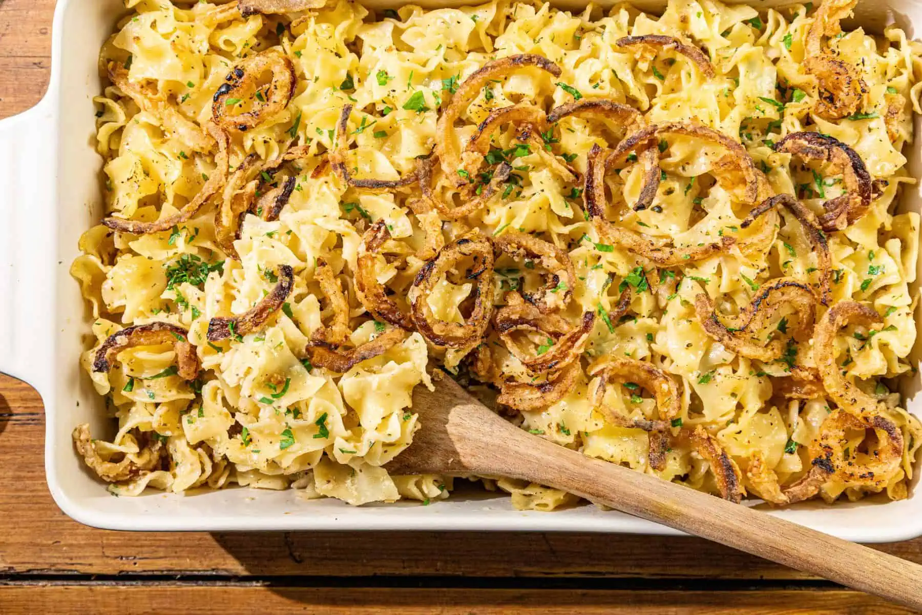 A French Onion Noodle Casserole filled with creamy egg noodles, topped with crispy fried onions and chopped parsley, sits invitingly in a dish with a wooden spoon resting inside.