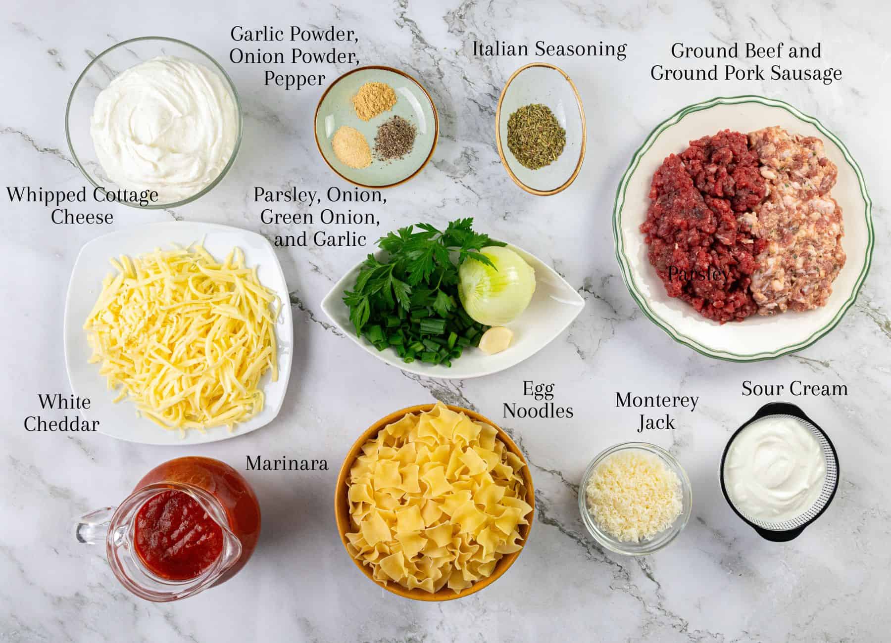 A marble surface displays shredded cheese, lasagna noodles, a jar of marinara sauce, portions of raw ground meat, small bowls with seasonings, and assorted fresh vegetables. Each item is labeled.