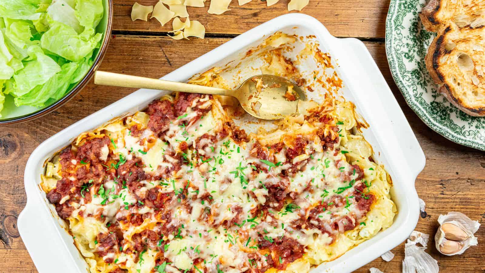 A baking dish of lasagna with melted cheese, a serving spoon, a bowl of lettuce leaves, toasted bread slices, and garlic on a wooden table.