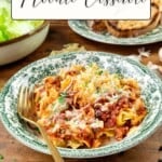 A plate of noodles mixed with beef and sour cream, topped with melted cheese and parsley, sits beside a bowl of leafy lettuce and a plate of toasted bread slices.