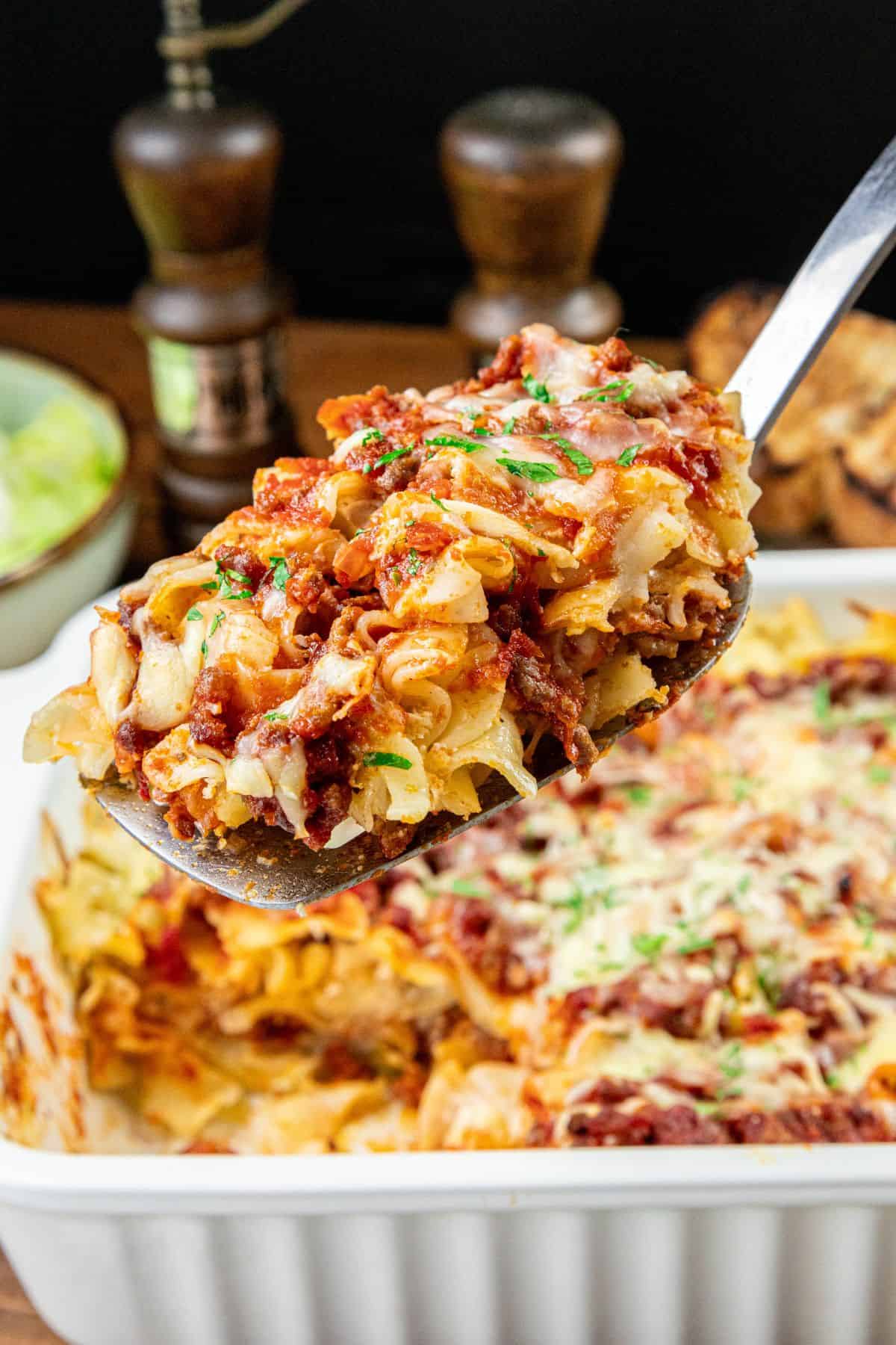 A serving spoon holds a portion of baked pasta covered in red sauce and melted cheese above a white casserole dish filled with more pasta.