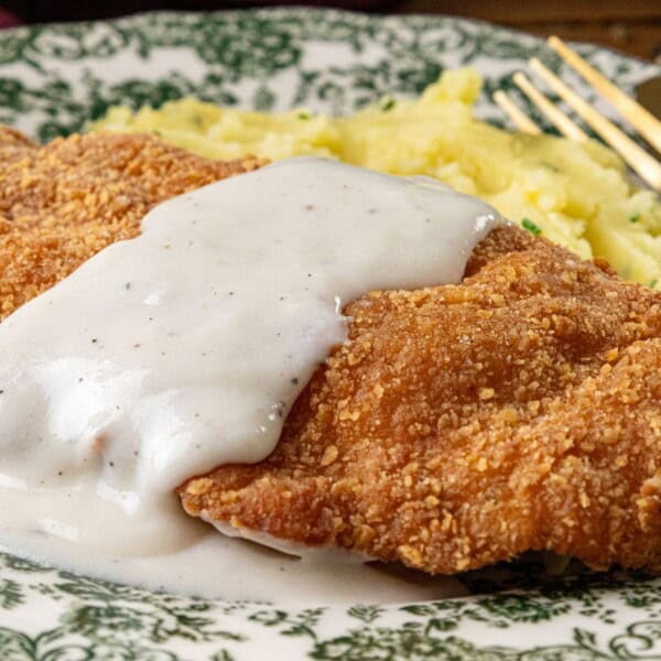 A plate with breaded fried chicken topped with white gravy, mashed potatoes on the side, and a fork and knife next to the plate.