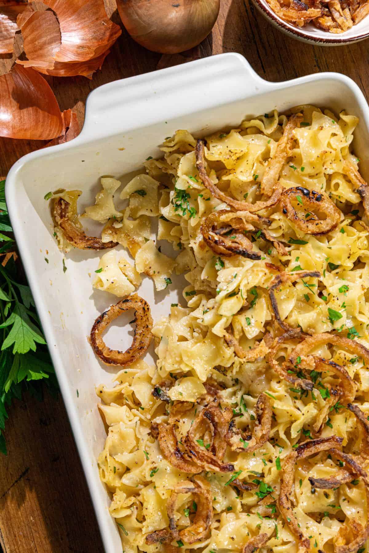 A white baking dish with a noodle casserole topped with crispy fried onions and chopped herbs, partially served, sits on a wooden surface beside onion skins and sprigs of fresh parsley.