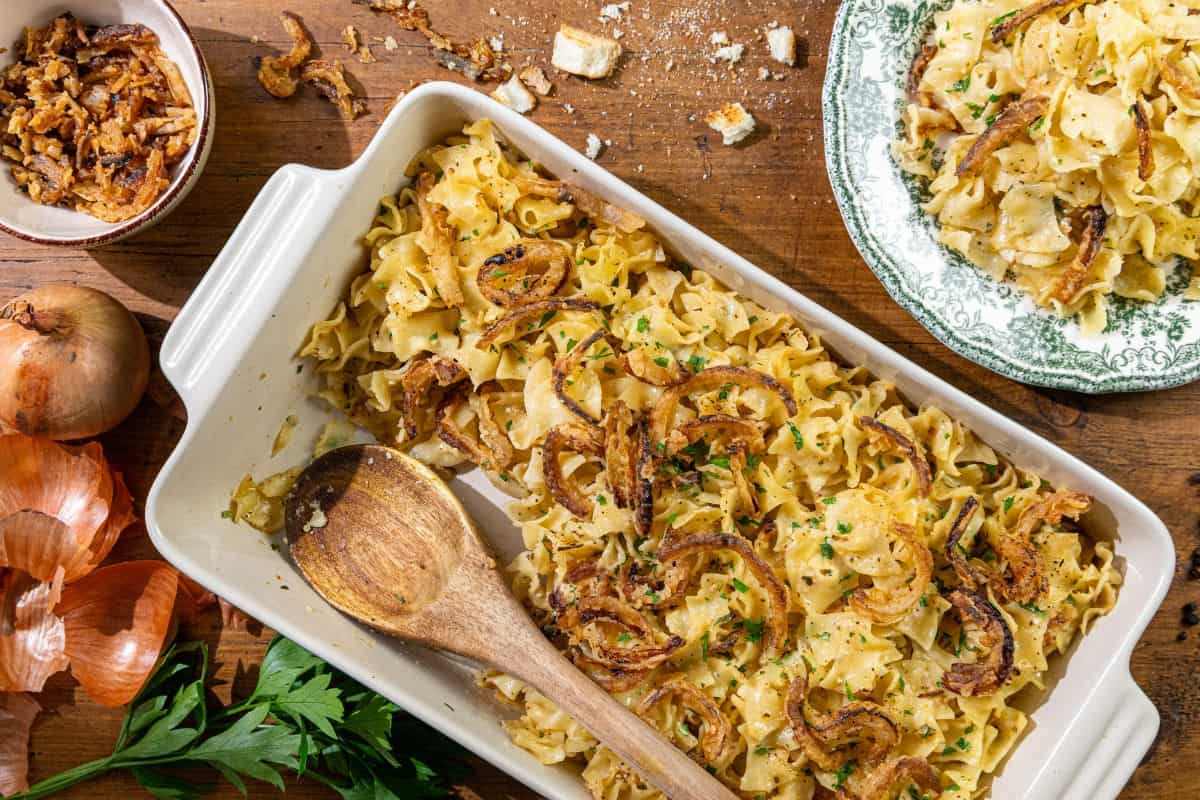 A baking dish filled with noodle casserole topped with fried onions, a wooden spoon, a bowl of extra fried onions, whole onions, fresh parsley, and a plate with a serving of the casserole.