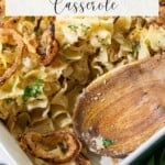 A casserole dish filled with noodles, topped with crispy onions and chopped herbs, with a wooden spoon resting inside.