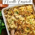 A casserole dish containing noodles topped with crispy onions and herbs, with a wooden serving spoon resting inside.