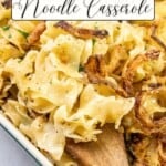 A white dish filled with French onion noodle casserole, topped with crispy onions, and a wooden spoon resting inside.
