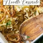 A white baking dish filled with noodle casserole, topped with crispy onions and chopped parsley. A wooden serving spoon rests in the dish.
