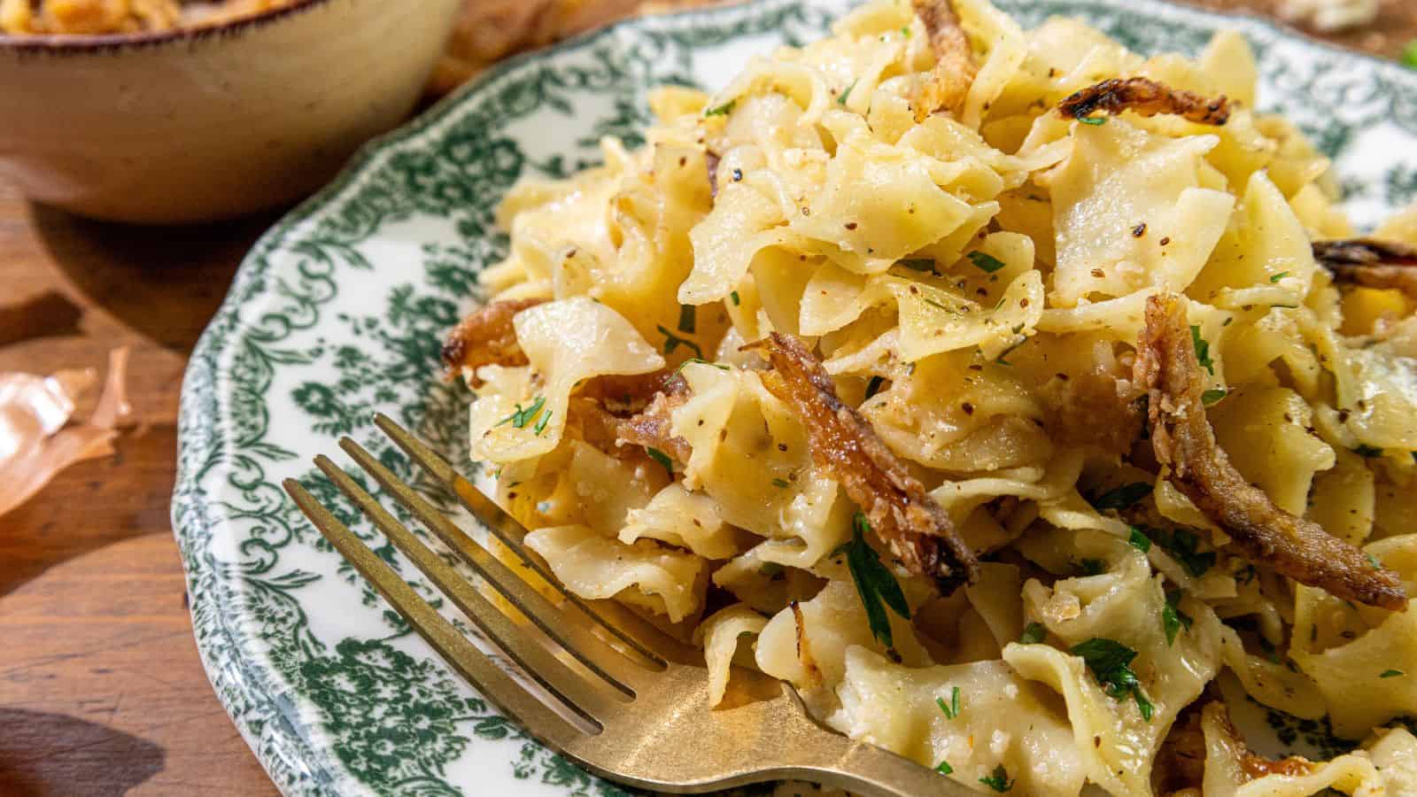 A plate of egg noodles topped with caramelized onions and herbs, accompanied by a fork and presented on a patterned dish.