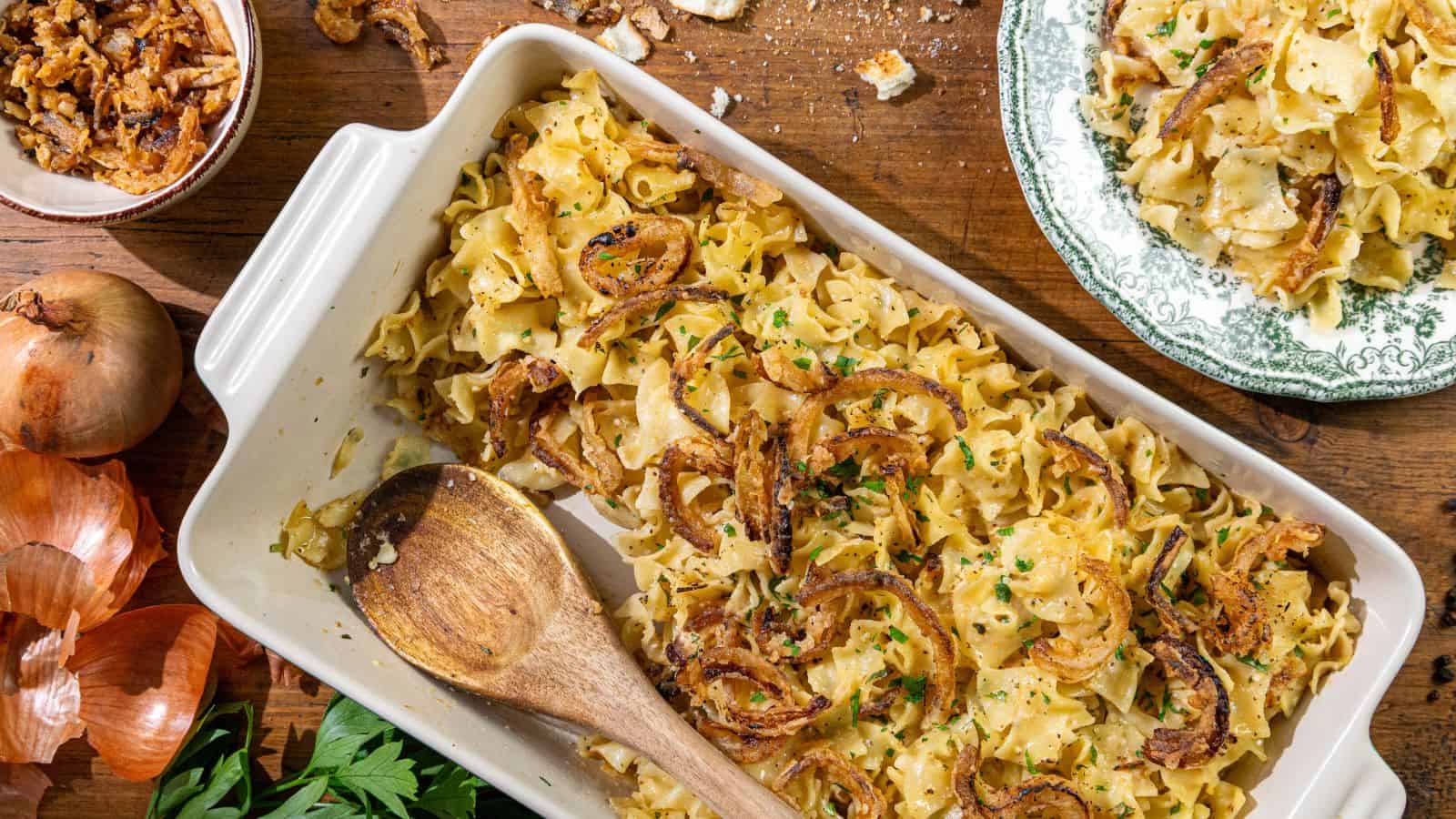 A casserole dish filled with cooked egg noodles, topped with crispy fried onions. A wooden spoon rests nearby, along with whole onions and a bowl of extra fried onions, all arranged on a wooden table.