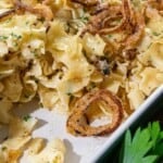 Cooked egg noodles mixed with herbs and topped with crispy fried onions, served in a white dish.