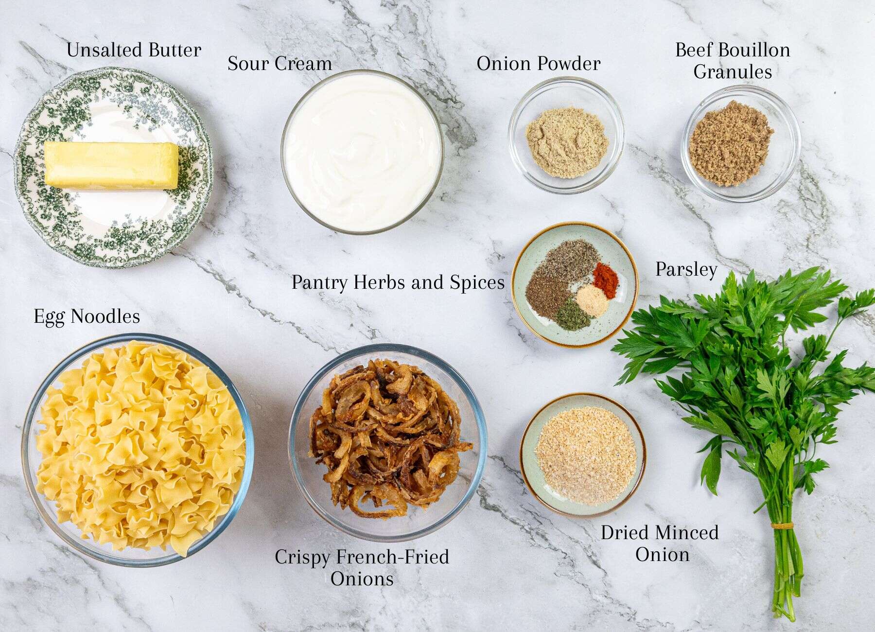 Butter, sour cream, uncooked egg noodles, crispy fried onions, bouillon cubes, dried herbs and spices in small bowls, and fresh parsley are arranged on a marble surface.