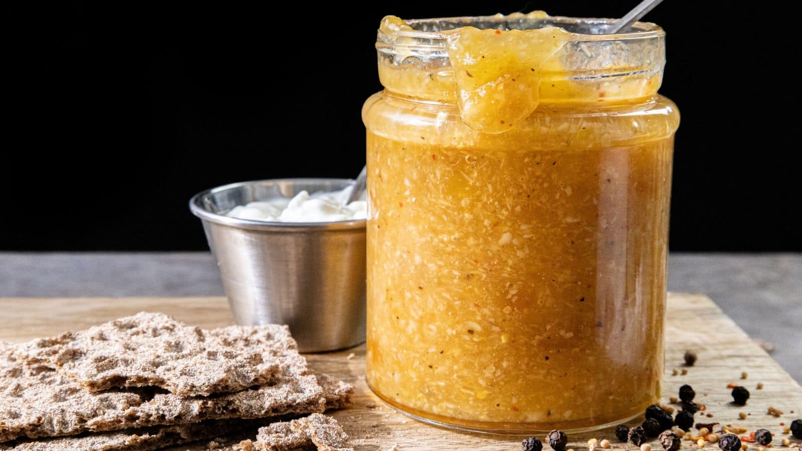 A jar filled with yellow relish containing a spoon, a metal cup holding white dip, several pieces of crispbread, and scattered peppercorns arranged on a wooden board.