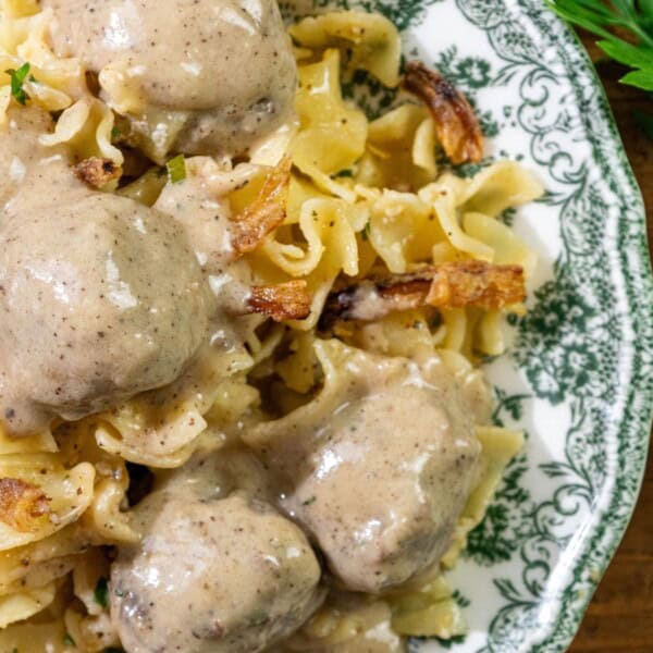 A plate of egg noodles topped with meatballs and creamy brown gravy, garnished with chopped parsley. Fresh parsley leaves are placed on the wooden table next to the plate.