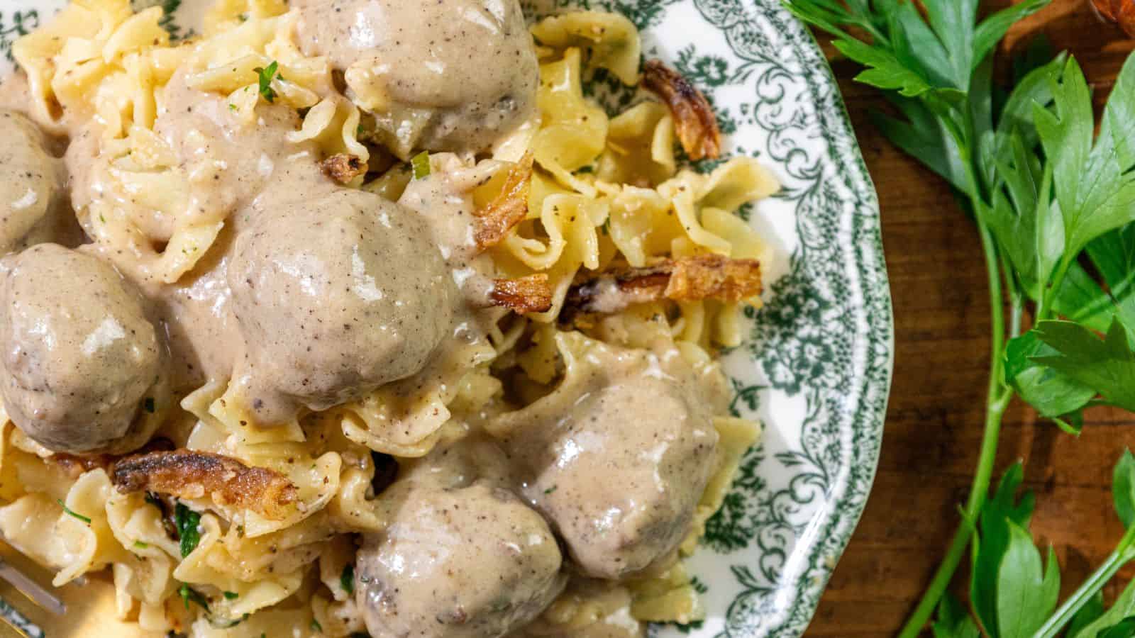 Egg noodles topped with meatballs and creamy brown sauce, garnished with chopped parsley, served on a plate beside sprigs of fresh parsley on a wooden table.