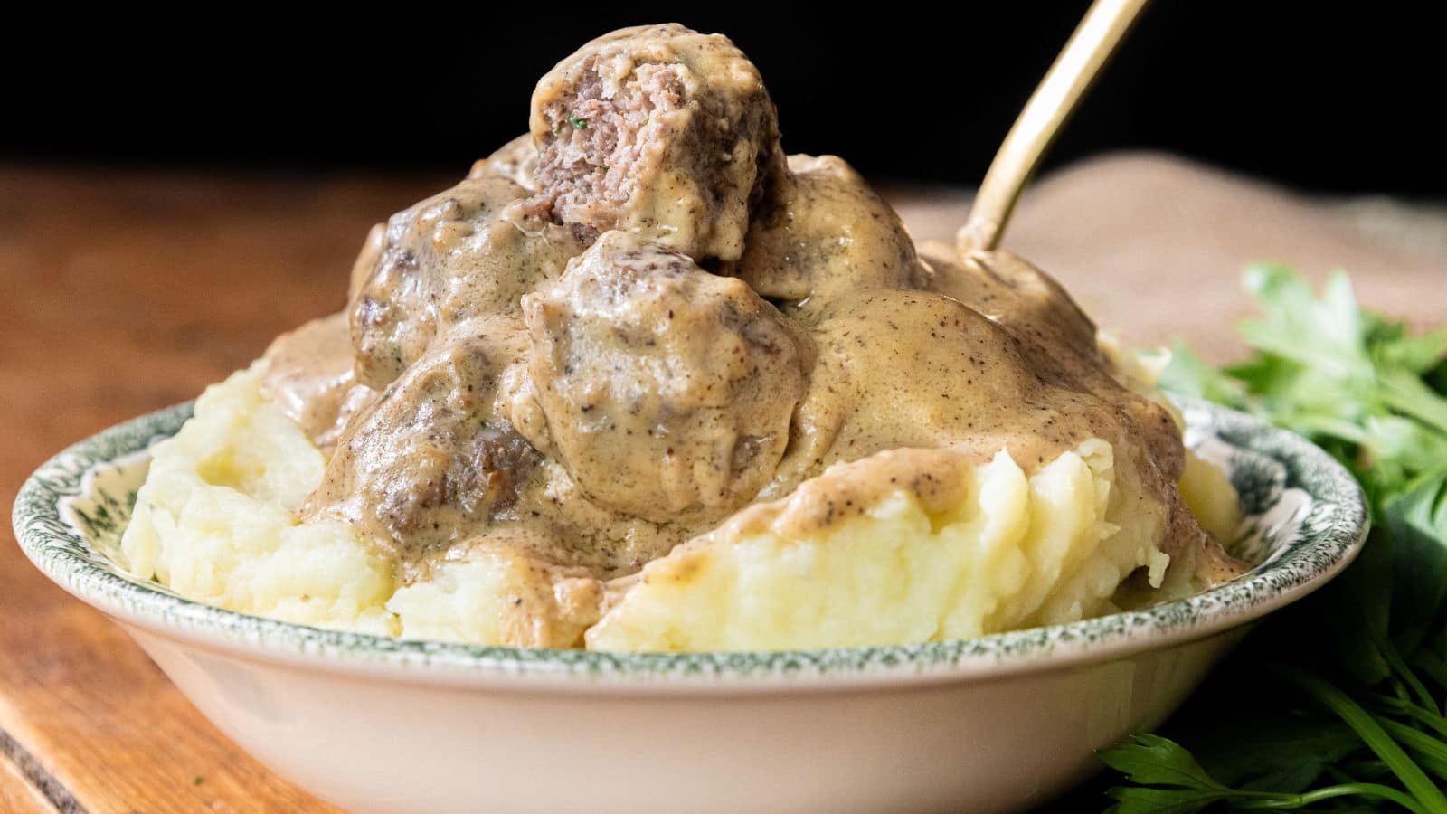 A bowl of mashed potatoes with meatballs covered in brown gravy and a spoon inside.
