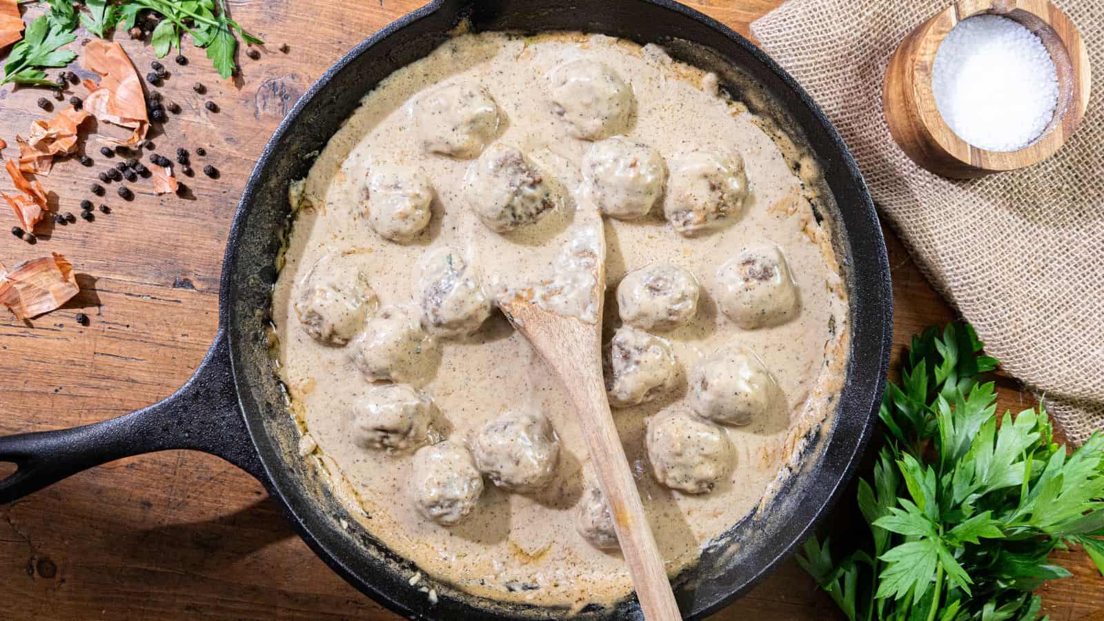 A cast iron skillet containing meatballs in a creamy sauce, garnished with fresh herbs, with a wooden spoon resting inside, placed on a wooden table.