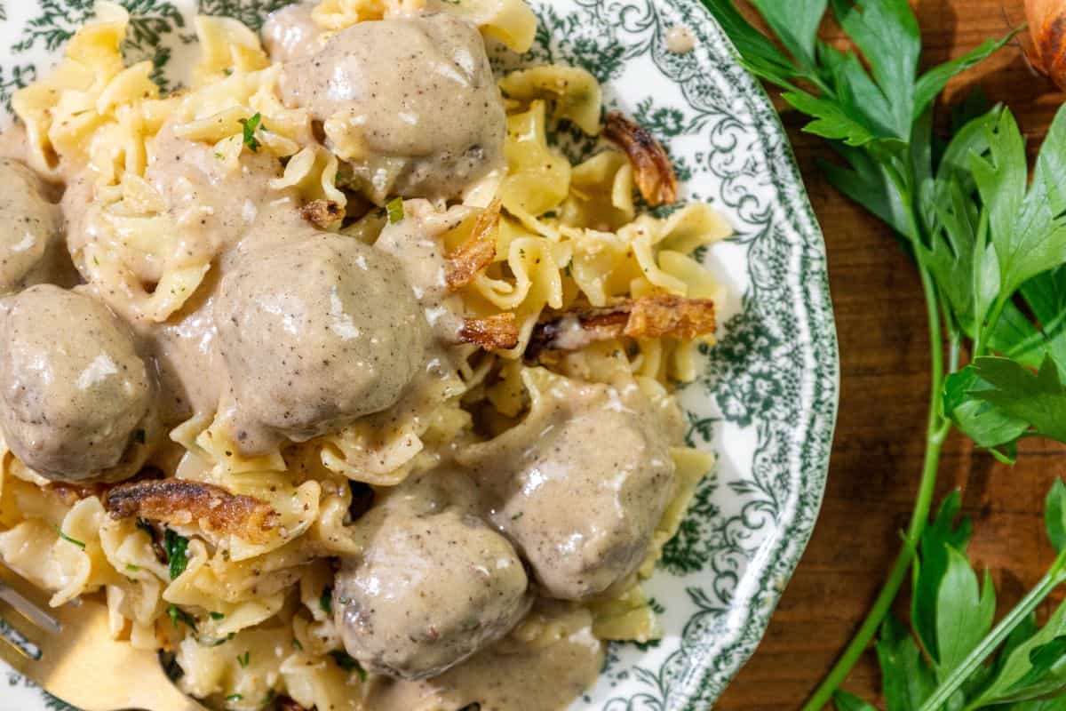 A patterned plate with pasta, meatballs, and creamy sauce is garnished with chopped parsley. Fresh parsley lies nearby on a wooden surface.