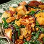 Sautéed beet greens topped with golden raisins, pine nuts, crispy croutons, and slices of prosciutto on a patterned plate, with a fork beside the food.