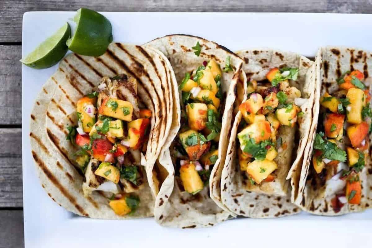 Three grilled tacos filled with diced vegetables and herbs are arranged on a white plate alongside two lime wedges.